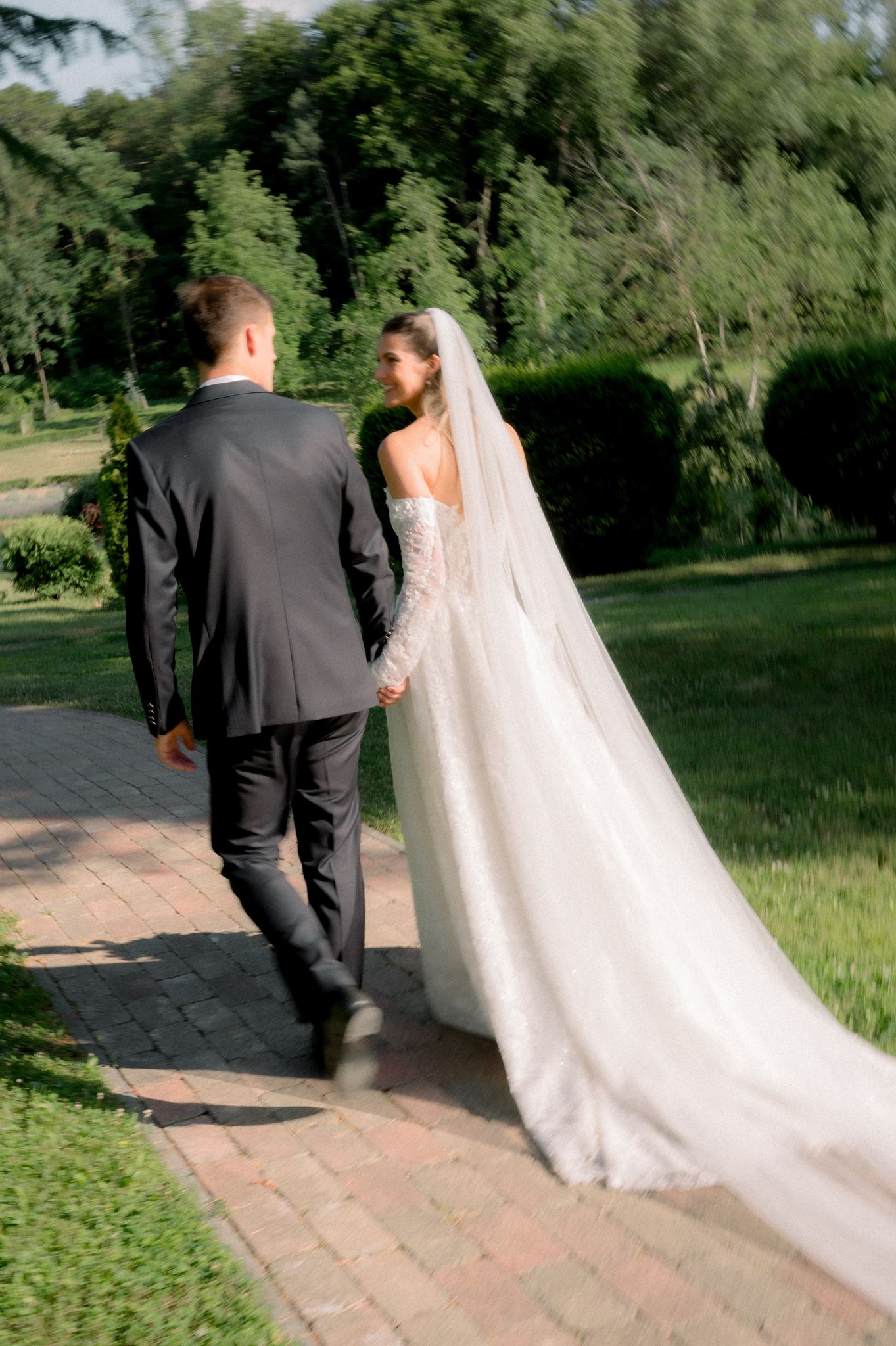 a couple in wedding attire walking away from the camera, looking and smiling at each other with green sorroundings